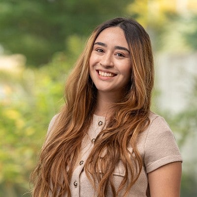 Headshot of Diana Santoyo smiling wearing light shirt against background of greenery.