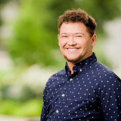 Headshot of Kason Ortiz-Carte smiling wearing a dark blue shirt against a blurred background of green plants.